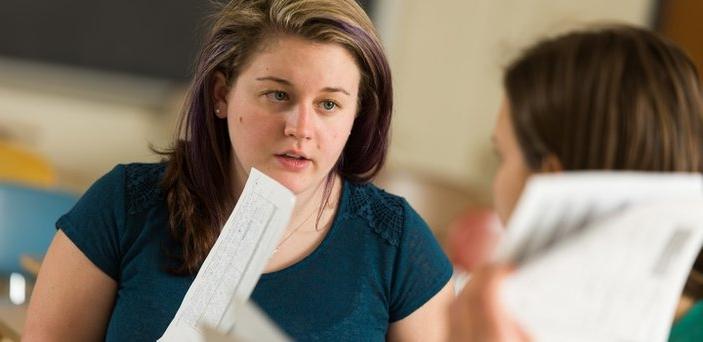 Two students at desks talking and holding coursework